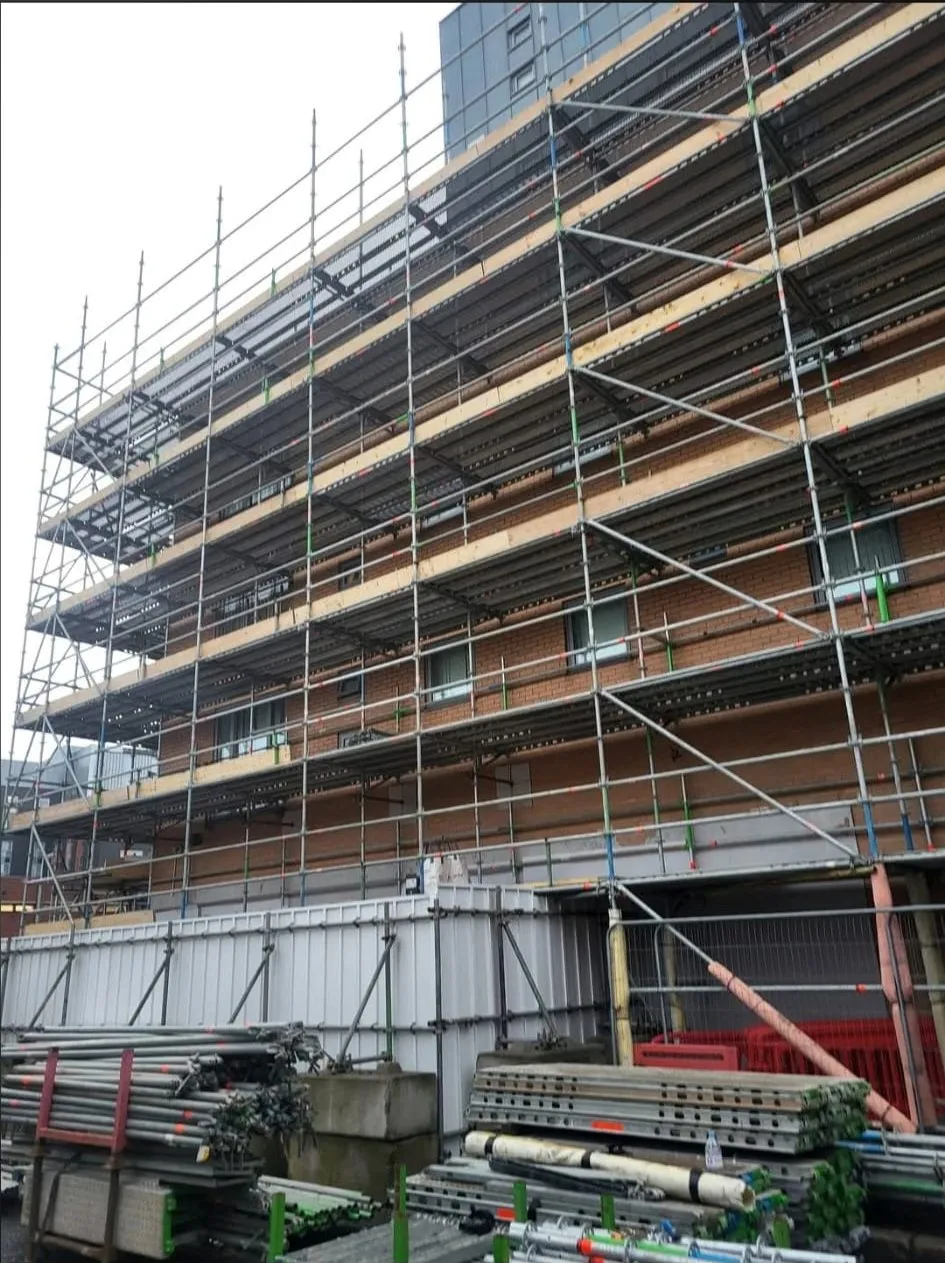Boarded scaffold across a commercial brick elevation with scaffold materials stacked at ground level
